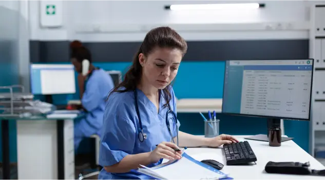 A woman in scrubs is seated at a desk, using a computer to process medical billing for therapists.